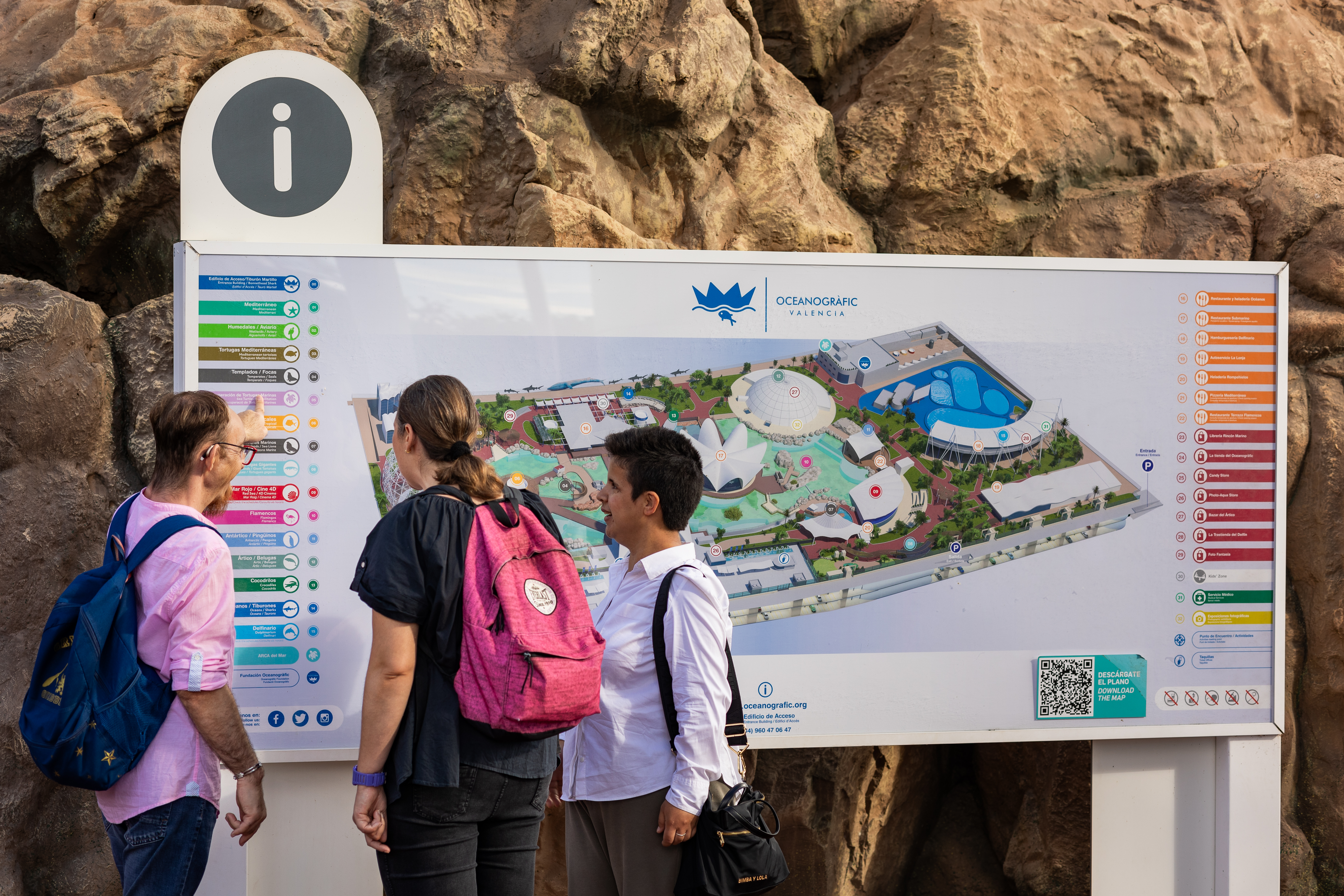 Tres personas, dos mujeres y un hombre, observan un gran plano informativo del Oceanogràfic de València instalado en el exterior. El panel muestra un mapa detallado del recinto con iconos de colores y una leyenda con pictogramas. La escena refleja accesibilidad e inclusión en el entorno turístico.