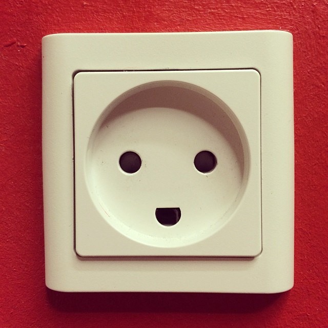 Fotografía de un enchufe con apariencia de cara sonriente en medio de una pared de color rojo.