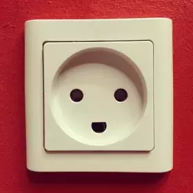 Fotografía de un enchufe con apariencia de cara sonriente en medio de una pared de color rojo. 